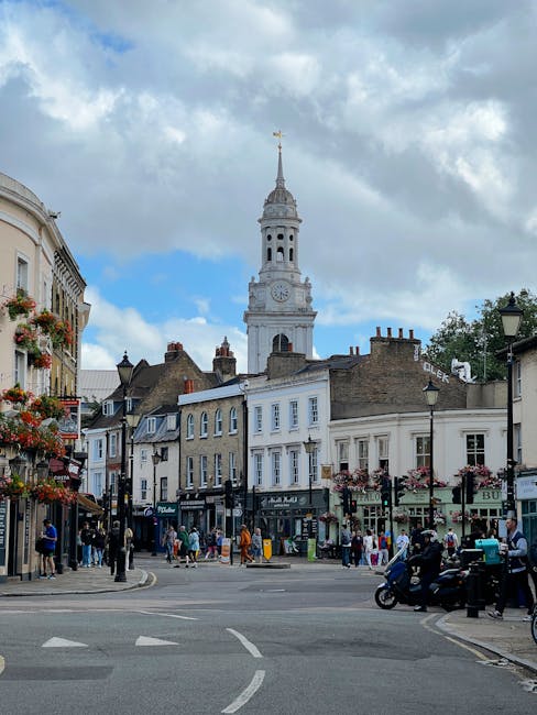 The image depicts a lively street scene in SW7 near Exhibition Road, featuring a prominent historic clock tower with a tall, ornate spire under a partly cloudy sky. Surrounding the tower are classic G