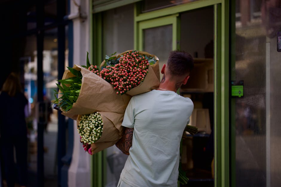 A man with short dark hair and a tattooed arm, wearing a light gray t-shirt, is seen from behind carrying a large bouquet of fresh flowers in brown paper wrapping. The bouquet features pink and white
