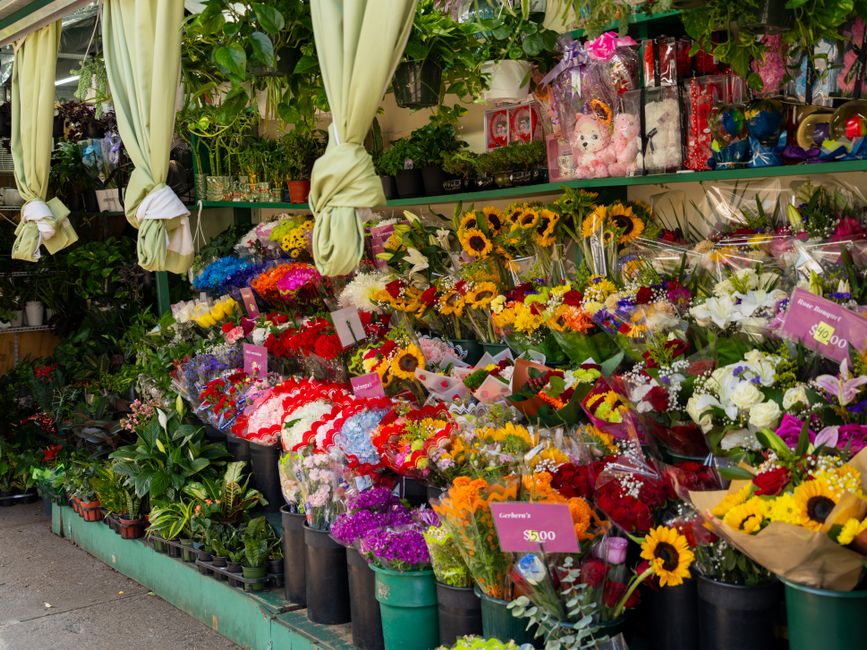 A vibrant floral display outside a flower shop features a variety of fresh flowers including yellow sunflowers, red roses, purple chrysanthemums, white lilies, and assorted mixed bouquets wrapped in c