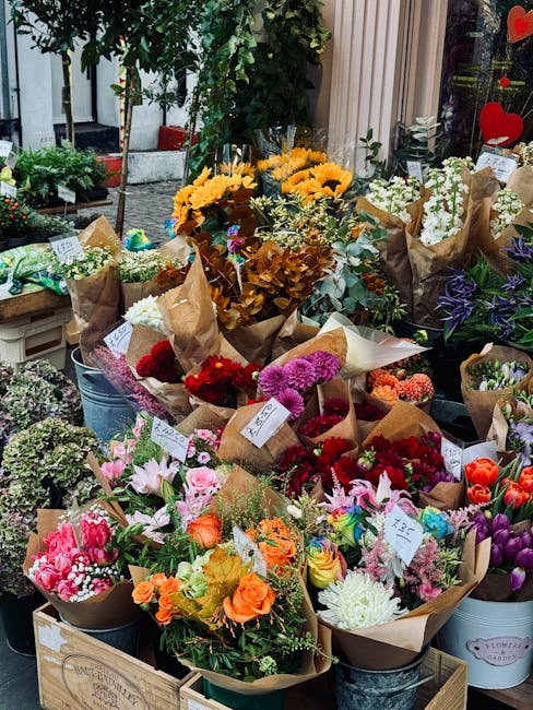 A display of fresh flower bouquets arranged in brown craft paper and placed in metal buckets and wooden crates. The bouquets feature a variety of flowers including vibrant roses in shades of orange, p