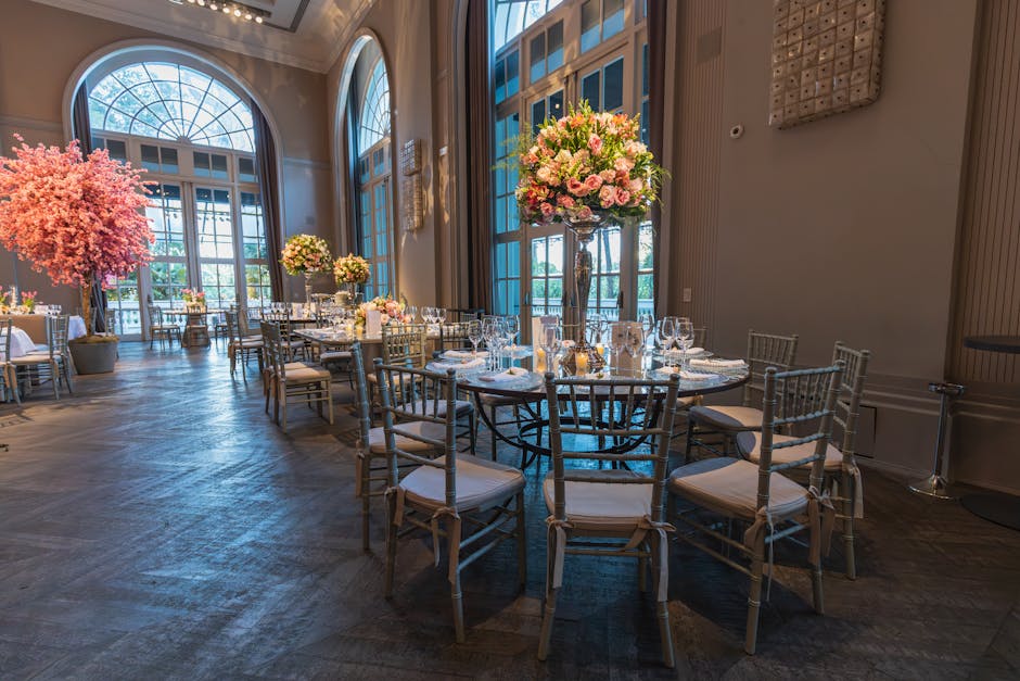 A spacious, elegantly decorated event room with high arched windows allowing natural light to fill the space. Central to the room are tall, clear glass vases with large, lush floral arrangements featu