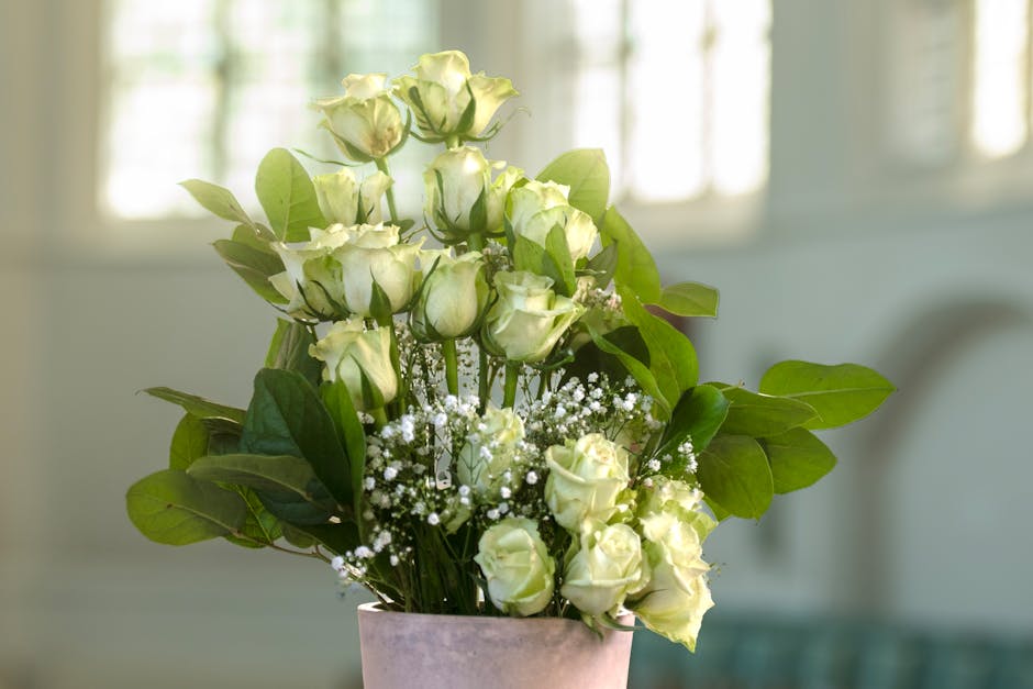 A bouquet of fresh, cream-colored roses with lush green leaves and small white baby's breath accents, arranged in a pink ceramic vase. The roses are in various stages of bloom, with some buds tightly 