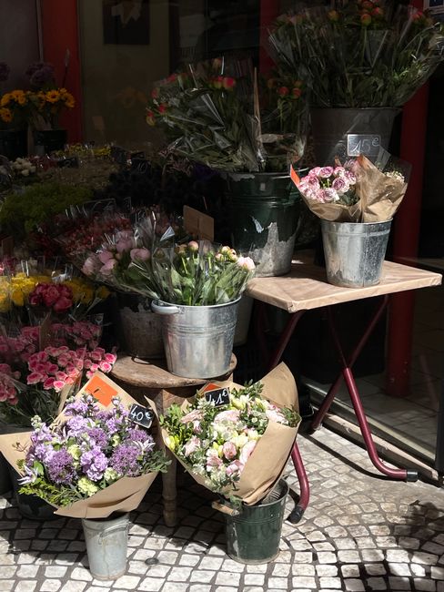 A display of freshly cut floral bouquets arranged in metal buckets and wrapped in brown paper, placed on small wooden and metal tables outside a flower shop. The bouquets include various flowers such