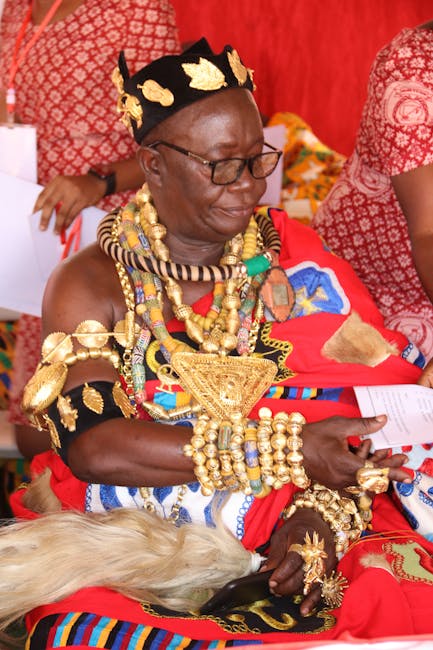 A woman dressed in traditional Maasai attire, wearing a vibrant red shuka and ornate jewelry made of gold, beads, and shells, seated indoors against a red patterned backdrop. She is adorned with layer