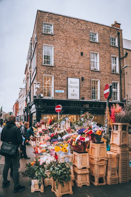 A bustling street scene in South Kensington featuring an outdoor flower market with numerous floral arrangements and bouquets displayed on wooden crates and tables. The flowers include a variety of co