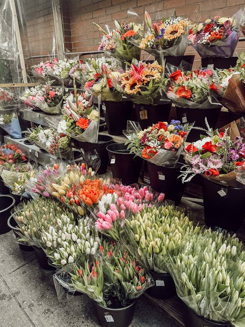 A large display of fresh flower bouquets arranged in black buckets and clear plastic wrapping, featuring a variety of flowers including pink, white, red, orange, purple, and peach tulips, roses, lilie