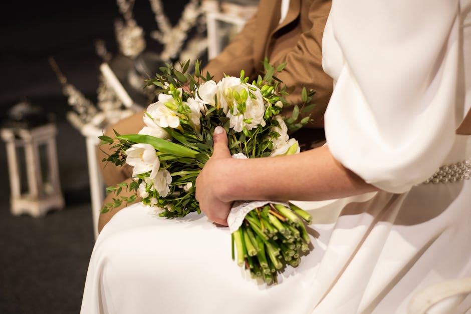 A close-up of a bride holding a bouquet of fresh white flowers, including lisianthus and greenery, with stems wrapped in white ribbon. She is seated, draped in a white wedding gown with a beaded belt,